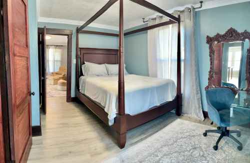 A cozy bedroom featuring a four-poster bed, a glass desk, and soft blue walls, with natural light streaming in.