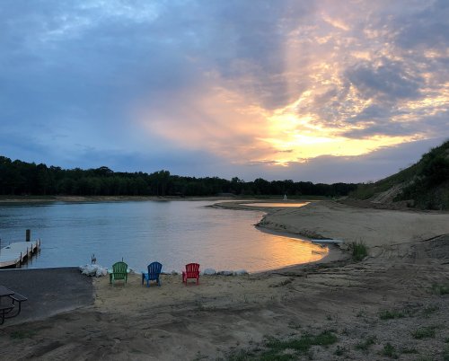 A serene lakeside scene at sunset, featuring colorful chairs on the sandy shore and a calm water surface.