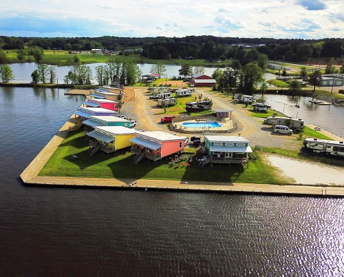 Aerial view of colorful cabins and RVs by a lake, with a pool and green landscape in the background.