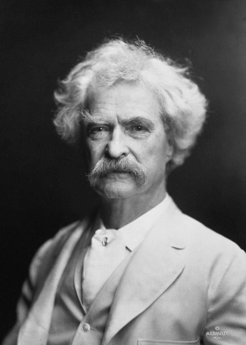 Black and white portrait of a man with a mustache and curly hair, wearing a light suit and looking directly at the camera.