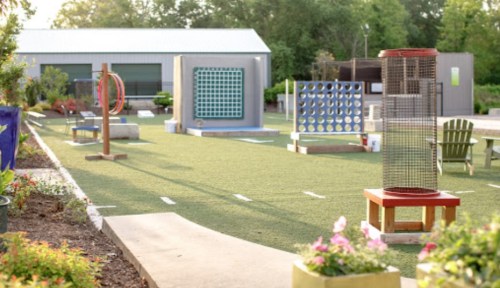 A vibrant outdoor play area with colorful games and activities on green turf, surrounded by trees and flowers.
