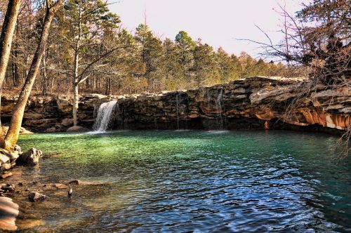 A serene natural scene featuring a waterfall cascading into a clear, greenish-blue pool surrounded by trees and rocks.