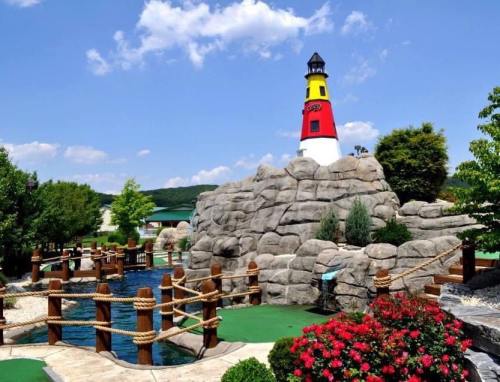 A colorful lighthouse atop a rocky formation, surrounded by a mini-golf course, water features, and vibrant flowers.