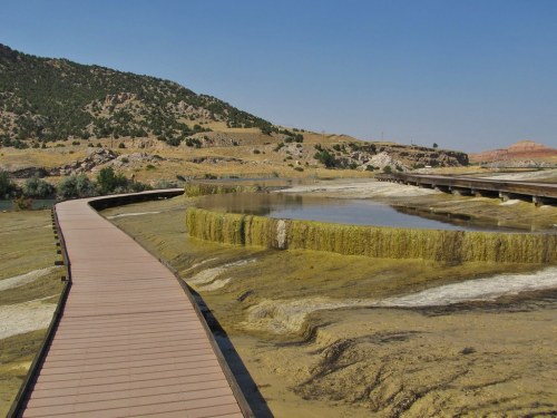A wooden walkway leads through a dry, rocky landscape with a small pond and grassy formations under a clear blue sky.