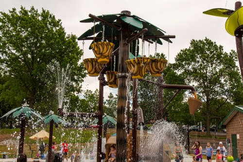 A water play area with colorful buckets and fountains, surrounded by trees and people enjoying the splash zone.