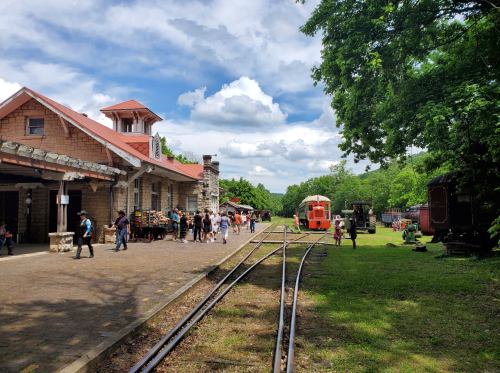 A historic train station with people gathered, surrounded by green trees and a vintage train on the tracks under a blue sky.