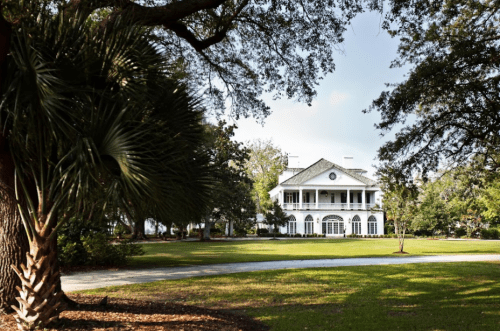 A grand white mansion with a large porch, surrounded by lush greenery and palm trees on a sunny day.