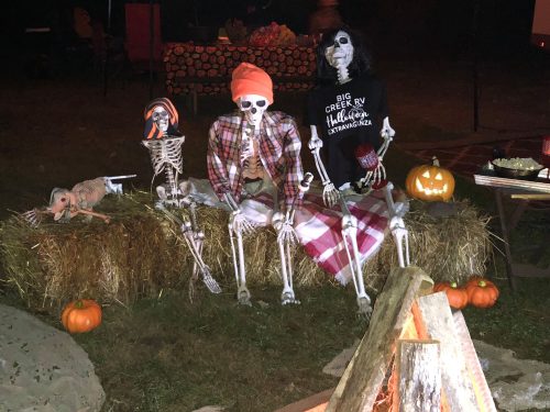 Three skeletons dressed in festive attire sit on hay bales by a campfire, surrounded by pumpkins and Halloween decorations.