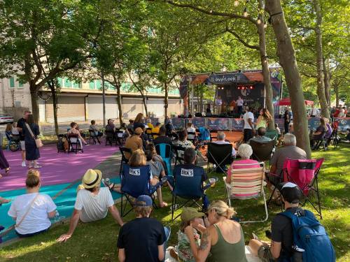 A crowd enjoys a live music performance outdoors, seated on the grass with colorful chairs and a stage in the background.