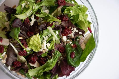 A fresh salad with mixed greens, dried cranberries, feta cheese, and sunflower seeds in a clear bowl.