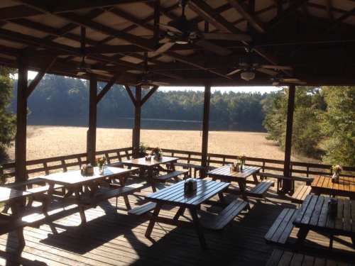 A rustic pavilion with wooden picnic tables overlooks a calm lake surrounded by trees. Sunlight filters through.