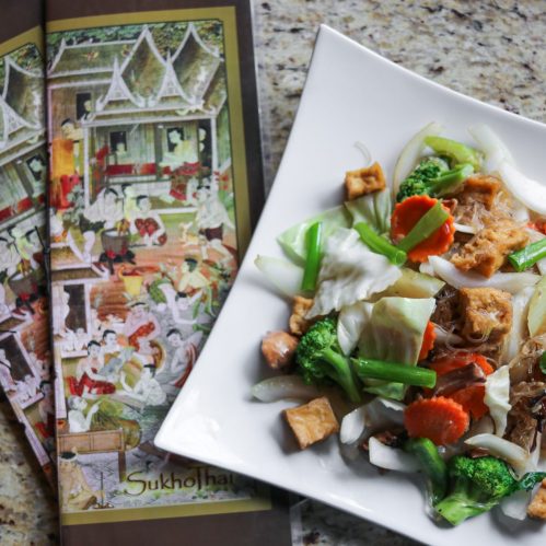 A plate of colorful stir-fried vegetables and tofu next to a book featuring traditional Thai art.