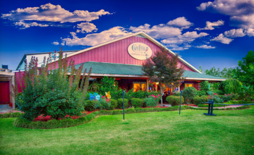 A colorful restaurant with a red exterior, surrounded by lush greenery and vibrant flowers under a blue sky with clouds.