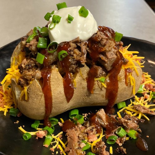 A loaded baked potato topped with shredded beef, cheese, barbecue sauce, green onions, and a dollop of sour cream.