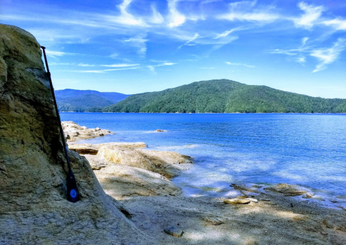 A serene lakeside view with rocky shorelines, blue water, and distant green hills under a clear sky.