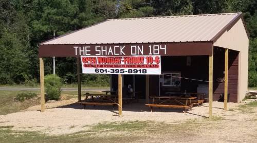 A small building with a sign reading "The Shack on 184," advertising hours and menu items, surrounded by trees.