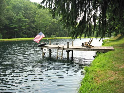 A wooden dock with an American flag, chairs, and a serene lake surrounded by trees.