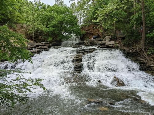 A cascading waterfall surrounded by lush green trees and rocky terrain, with people visible in the background.