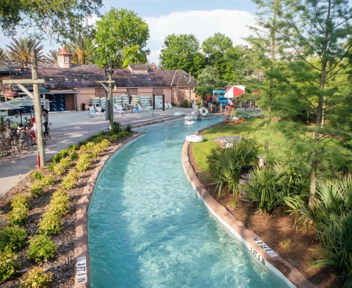 A winding lazy river surrounded by greenery, with lounge chairs and umbrellas in a sunny outdoor setting.