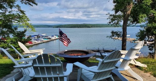A serene lakeside view with white adirondack chairs, a fire pit, and an American flag, surrounded by trees and boats.