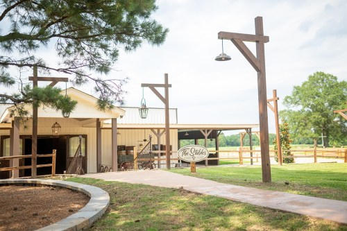 A rustic venue with wooden structures, a pathway, and a sign reading "The Stables," surrounded by greenery.