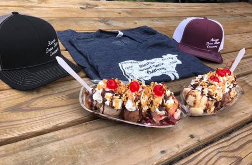 Two ice cream sundaes topped with nuts and cherries, alongside a t-shirt and two hats on a wooden table.
