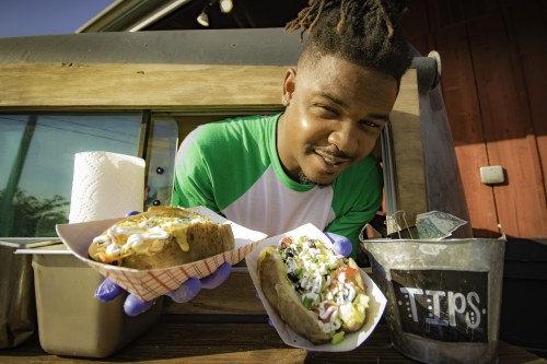 A smiling vendor holds two loaded hot dogs in front of a food truck, with a tip jar nearby.