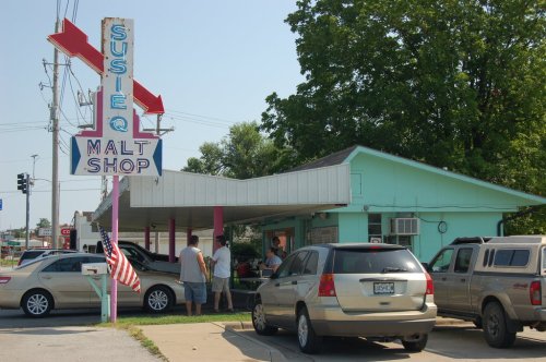 A colorful malt shop with a retro sign, surrounded by parked cars and trees on a sunny day.