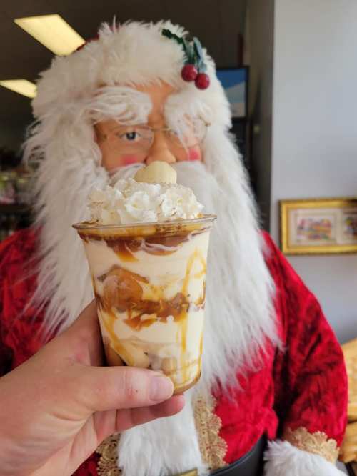 A hand holds a dessert cup with whipped cream and caramel, with a Santa figure in the background.