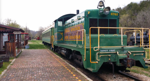 A green vintage train engine with "Eureka Springs" lettering parked on a brick platform surrounded by trees.