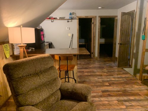 Cozy interior of a room with a recliner, bar area, and two doorways leading to other spaces. Warm lighting and wooden floors.