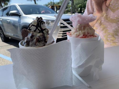 Two cups of ice cream topped with chocolate sauce and cotton candy, sitting on a table with a car in the background.