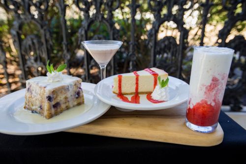 A wooden platter with a slice of cheesecake, a dessert with sauce, and a colorful drink, all garnished with whipped cream.