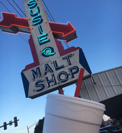 A retro neon sign for "Susie Q Malt Shop" with a drink in the foreground against a clear blue sky.