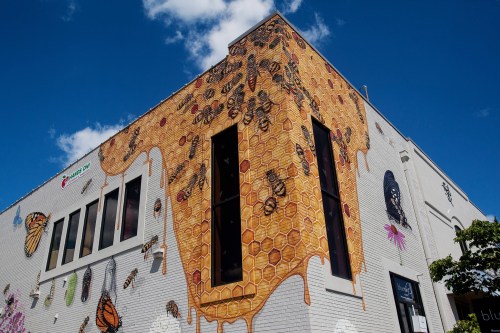 Colorful mural on a building featuring honeycomb, bees, and butterflies against a blue sky.