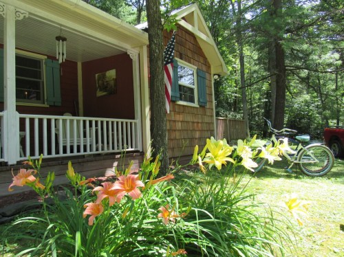 A cozy cottage with a porch, surrounded by colorful flowers and a bicycle nearby, set in a lush green landscape.