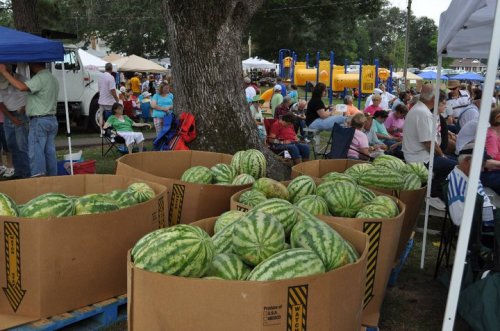 A bustling outdoor festival scene with large boxes of watermelons in the foreground and crowds enjoying the event.