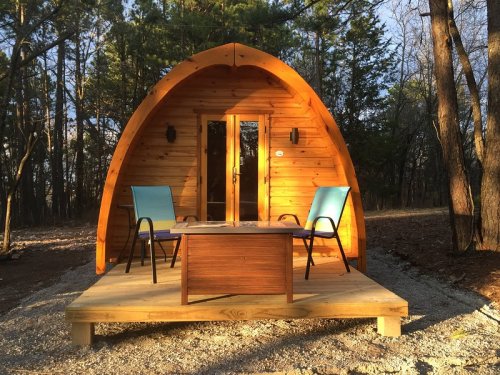 A cozy wooden cabin with an arched roof, surrounded by trees, featuring a small deck and two chairs.