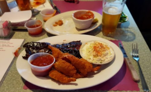 A plate of fried shrimp, ribs with barbecue sauce, coleslaw, and dipping sauces, with a beer on the side.