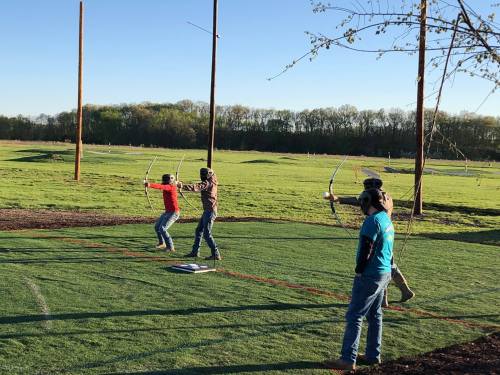 Four individuals practice archery on a grassy field, aiming at targets in the distance under a clear blue sky.