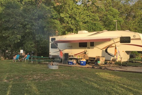 A camper parked on grass with chairs, a table, and a fire pit nearby, surrounded by trees in a sunny setting.