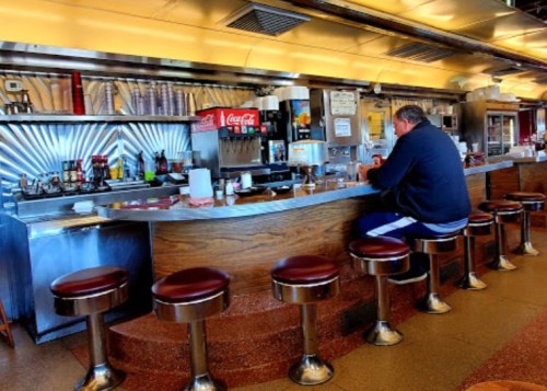 A diner interior with a man sitting at the counter, surrounded by empty stools and a retro decor.