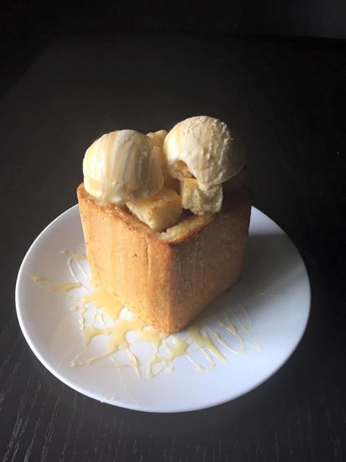 A square slice of bread topped with two scoops of vanilla ice cream and drizzled with syrup on a white plate.