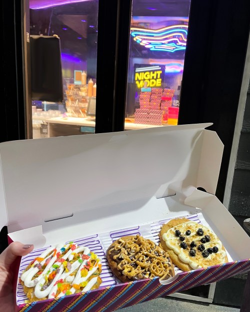 A hand holding a box of three decorated cookies in front of a brightly lit shop window with "NIGHT MODE" sign.