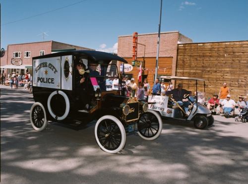 A vintage police car and a golf cart in a parade, with spectators watching from the sidelines on a sunny day.
