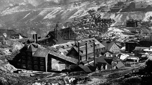 Historic black-and-white photo of a mining town with industrial buildings and mountainous terrain in the background.