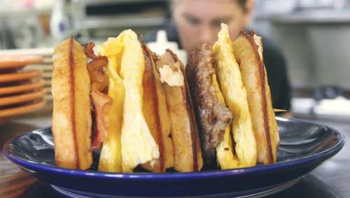 A plate with three sandwich halves, each filled with eggs, bacon, and sausage, set against a blurred kitchen background.