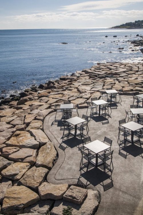Outdoor seating area with tables on a rocky shoreline, overlooking a calm sea under a clear sky.