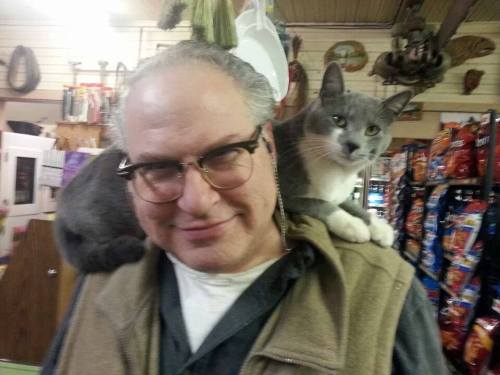 A man with glasses smiles while a gray cat perches on his shoulder in a store filled with snacks.