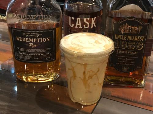 A cup with a creamy drink sits in front of three whiskey bottles on a wooden surface.